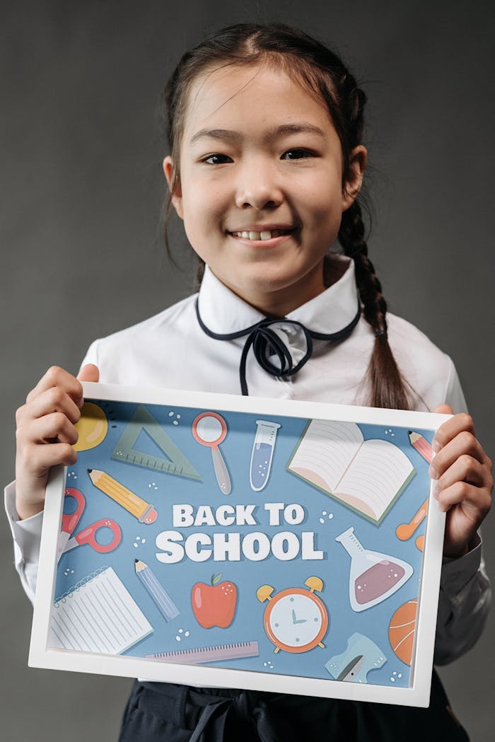 A young girl in a school uniform holds a back to school poster with a playful design.